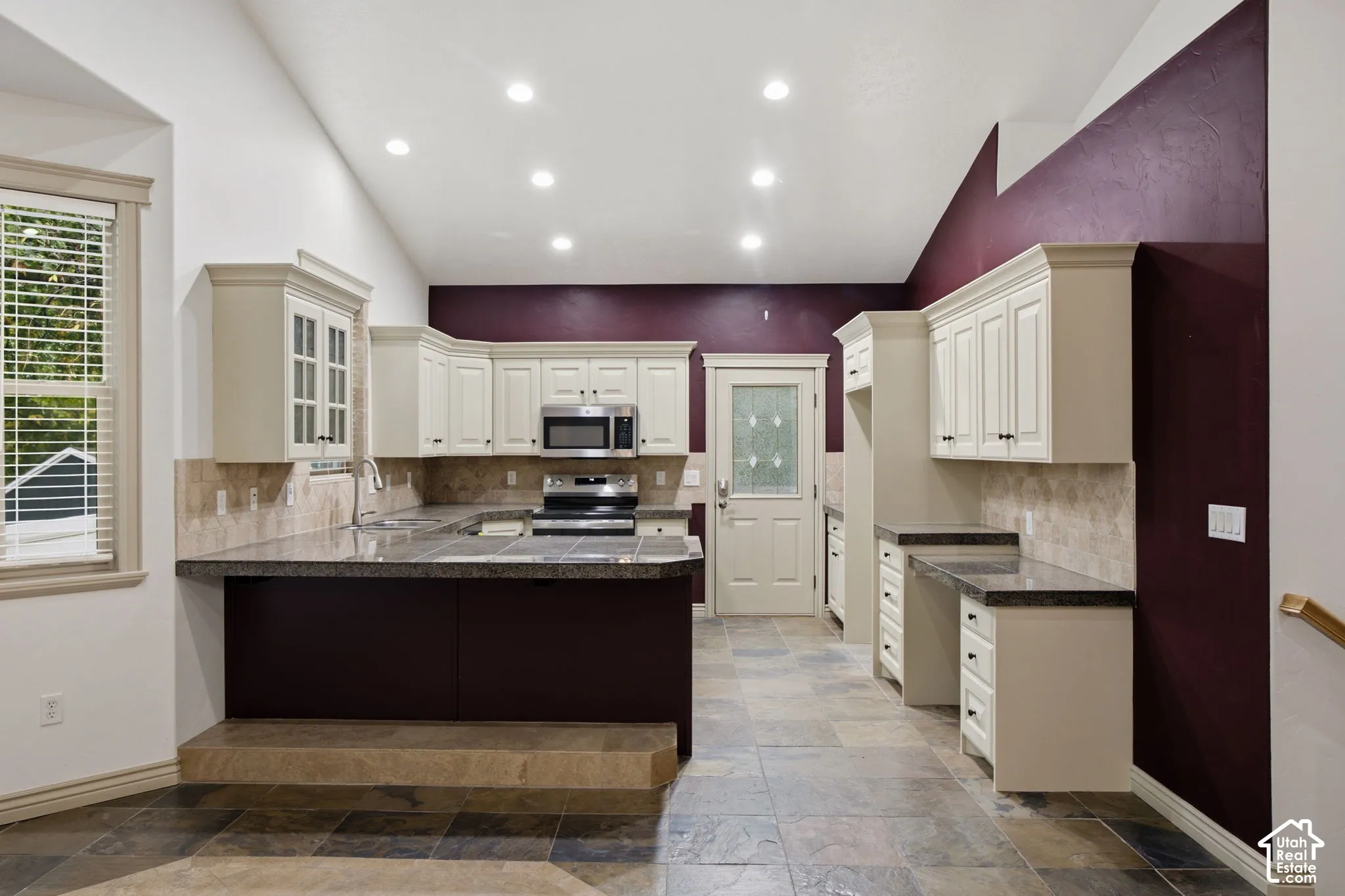 Kitchen featuring vaulted ceilings, light-tone cabinetry, stone-finish tile flooring, a tiered breakfast bar, and stainless steel appliances