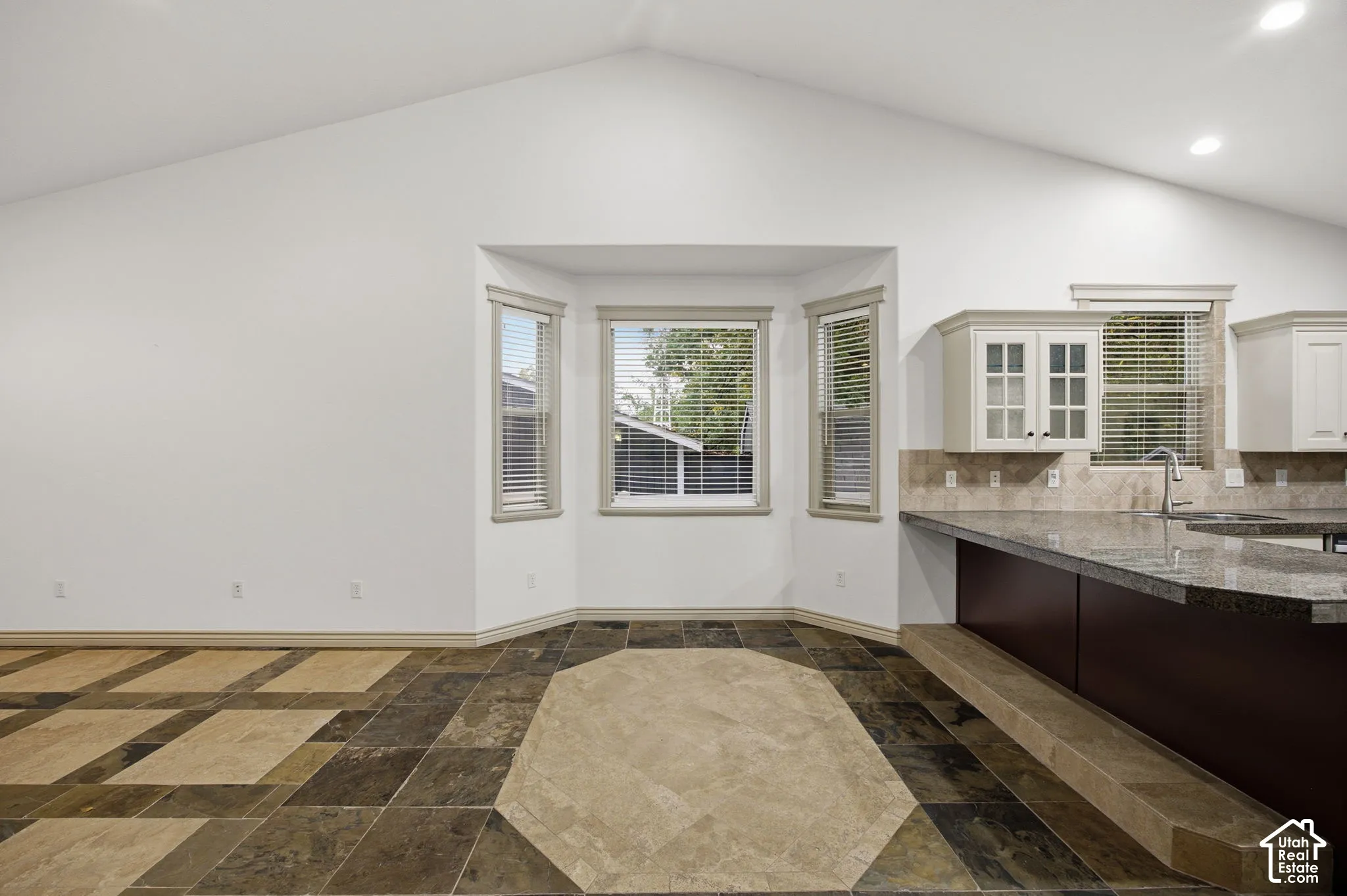 Vaulted ceilings, bay window, stone-patterned tile flooring, breakfast bar with stone countertop, and upper cabinetry with glass-panel doors