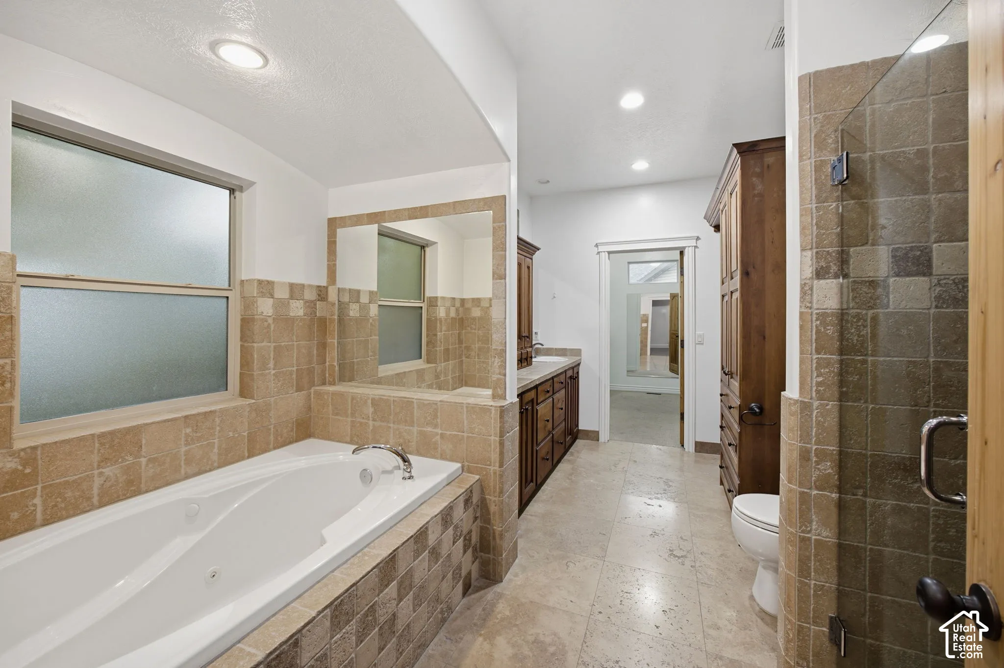 Bathroom featuring a jetted soaking tub, walk-in glass-enclosed shower, custom wood cabinetry, natural stone tile flooring, and wall tile surround