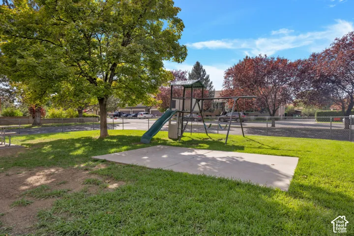 Outdoor recreation area featuring a concrete pad, metal play structure with slide, perimeter chain-link fencing, and mature trees