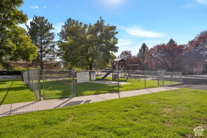 Fenced recreational area featuring a play structure, gravel surfacing, mature trees, and concrete perimeter walkway