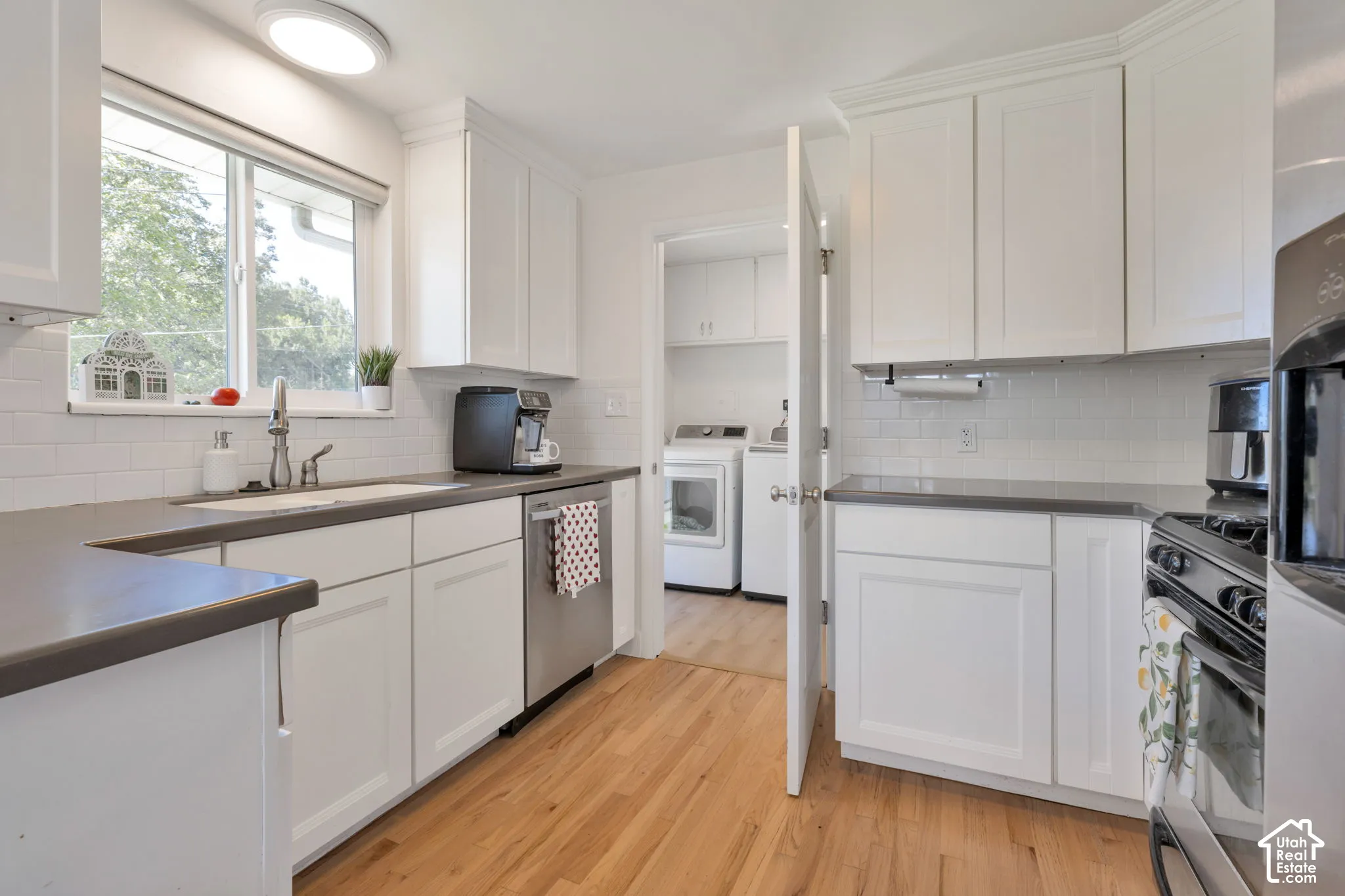 U[dated kitchen featuring white shaker-style cabinetry, solid-surface countertops, subway tile backsplash, stainless steel dishwasher, and wood-flooring