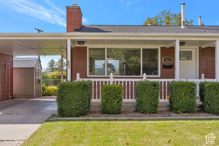 Single-story brick exterior featuring a covered front porch with white railings, attached carport, red brick chimney, and concrete walkway