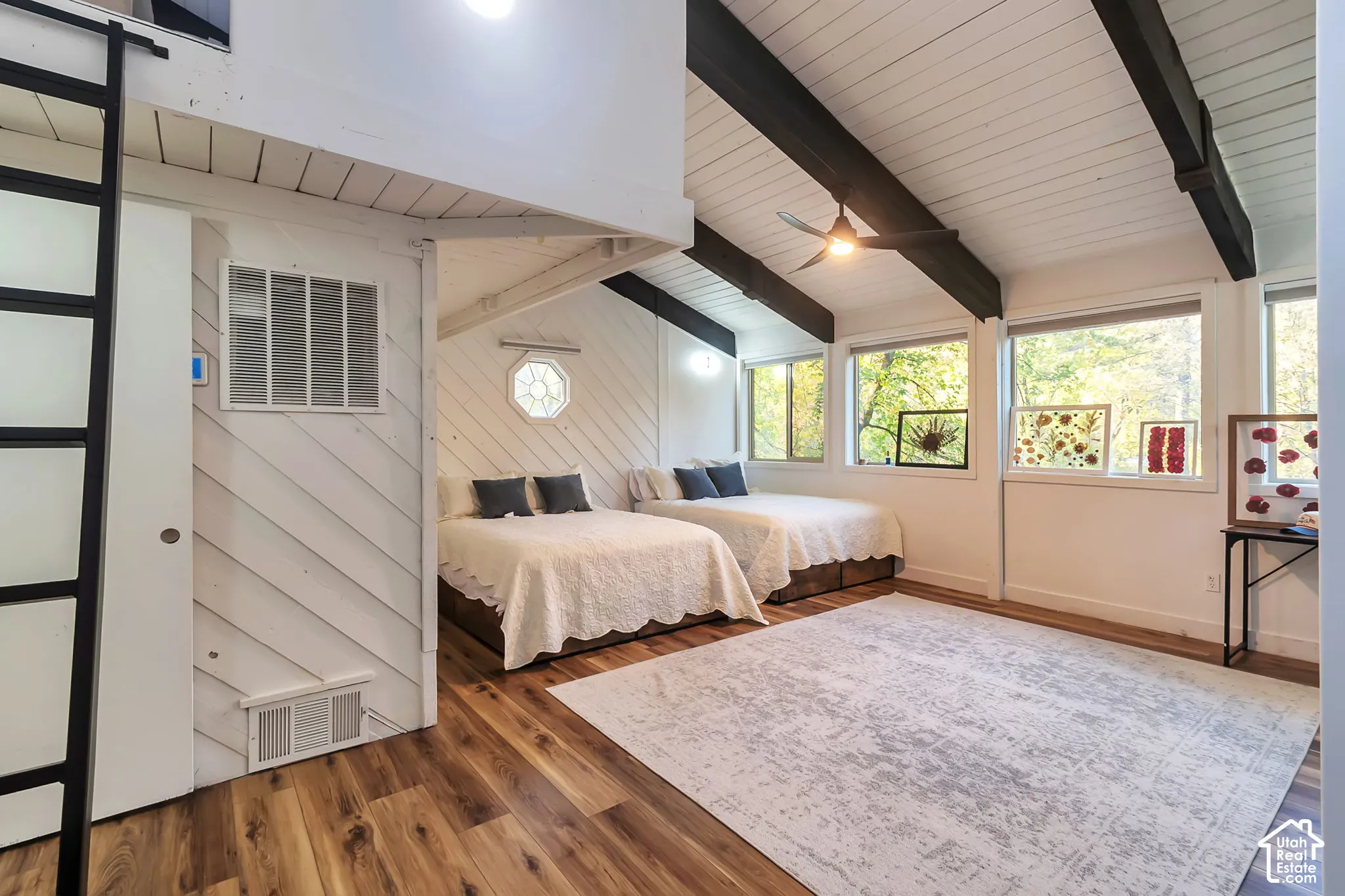 Vaulted ceiling with exposed dark-toned structural beams, white wood-finish wall paneling in a diagonal pattern, wood-finish flooring, and a built-in fixed ladder