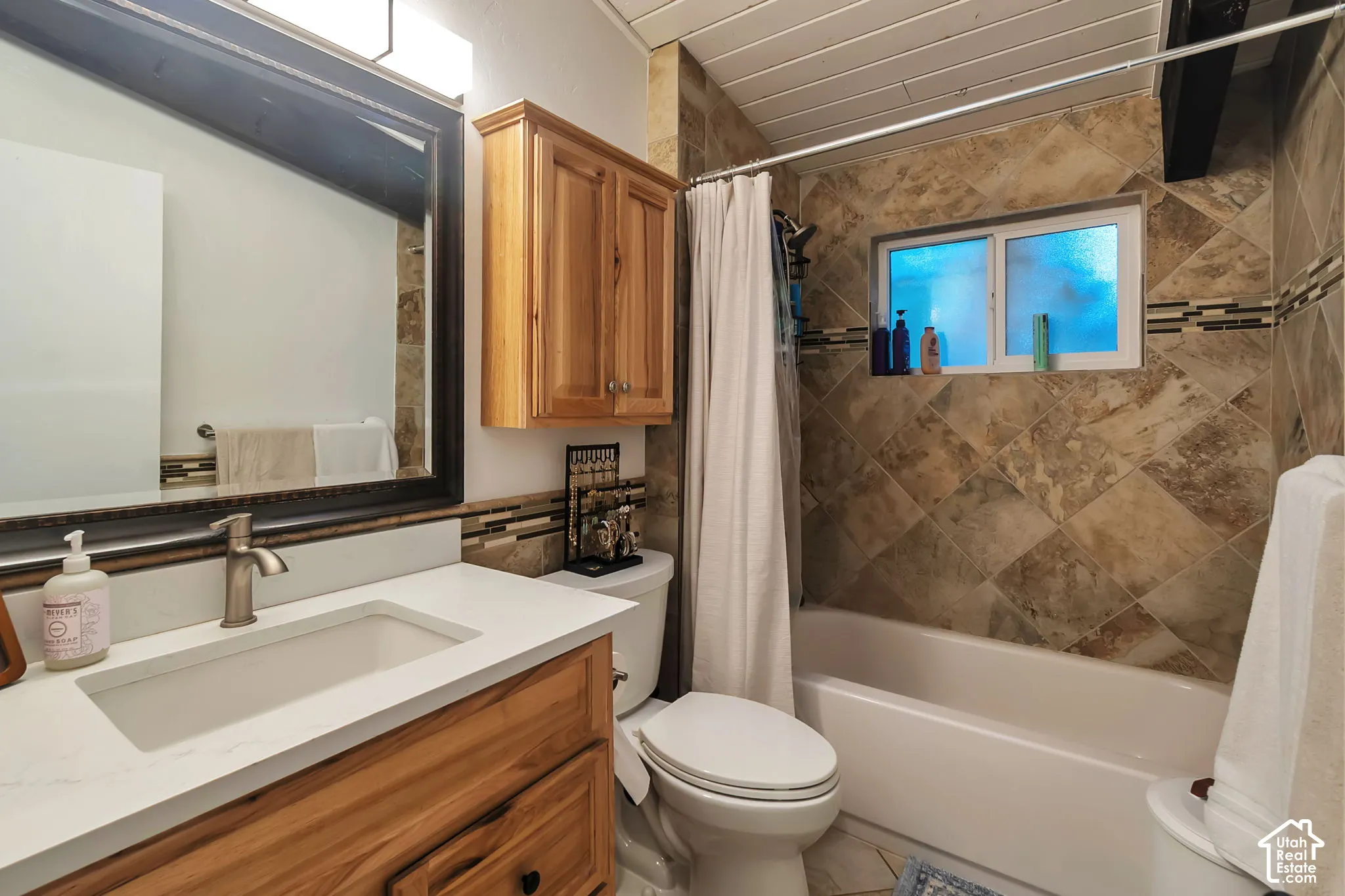 Bathroom featuring a vanity with a light wood finish, white countertop, integrated rectangular sink, wall-mounted wood cabinetry, and a bathtub surrounded by diagonal-set stone-style tiling