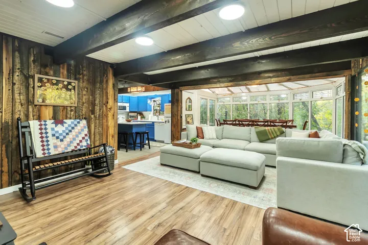 Living area featuring dark-stained exposed ceiling beams, vertical wood-paneled walls, wood-finish flooring, and a glass-enclosed sunroom