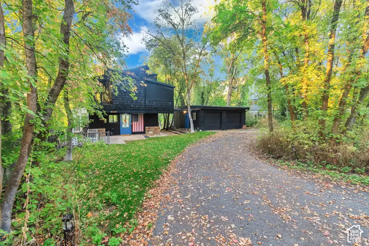 Two-story residence featuring dark shingle siding, a paved driveway, an attached garage, and stone masonry exterior accents surrounded by mature trees