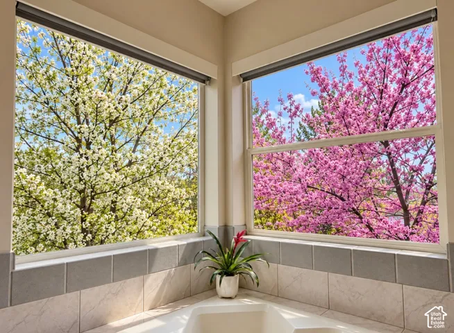 Corner windows featuring expansive glass panes, neutral tile wall surround, built-in soaking tub, and exterior botanical foliage