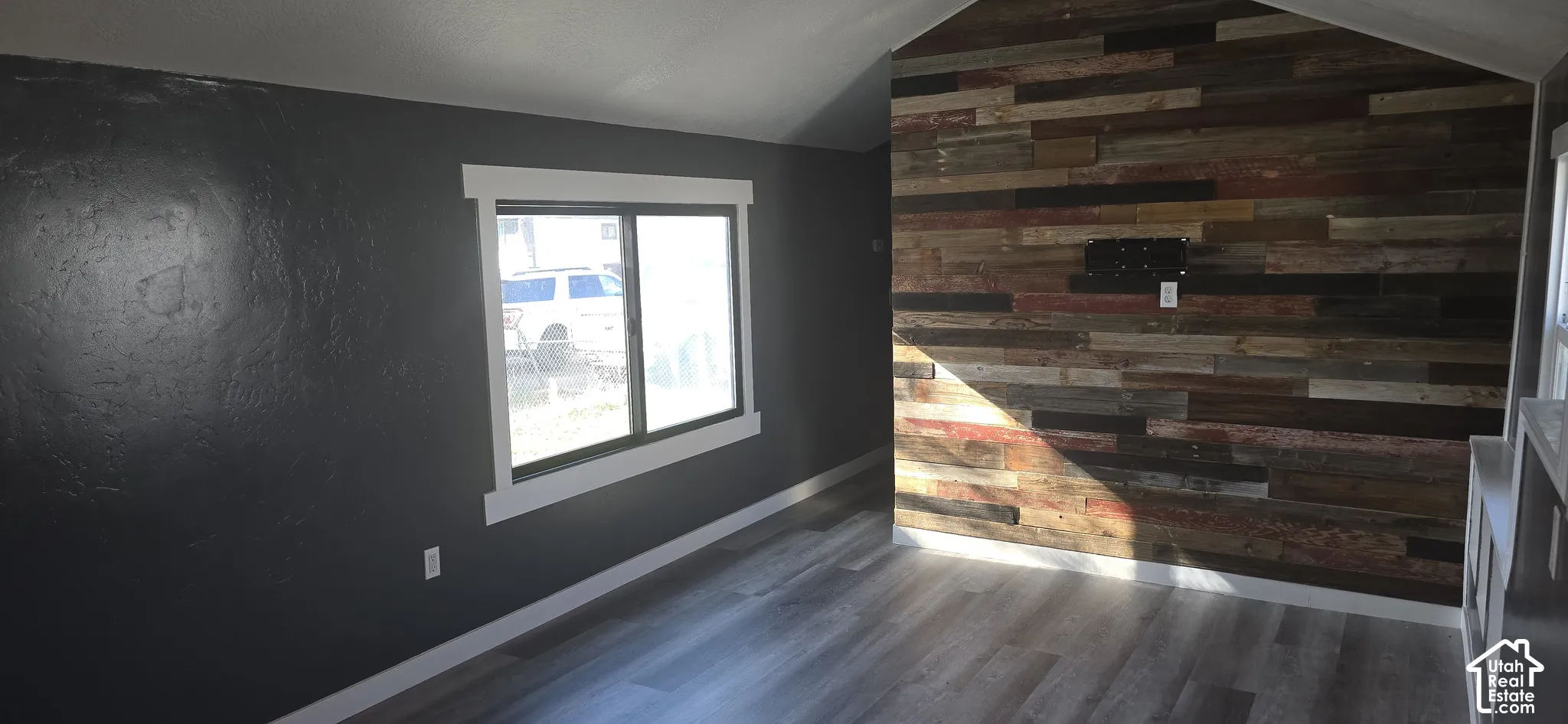 Vaulted ceiling featuring a reclaimed wood accent wall, wood-finish flooring, dark-toned walls, and thick white window trim