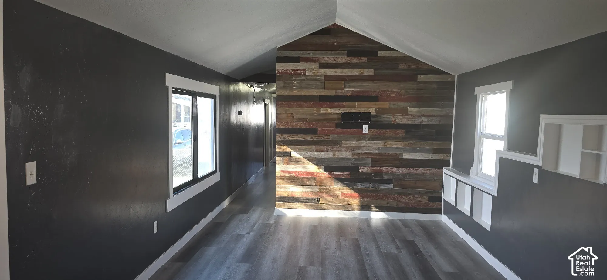 Vaulted ceiling room featuring a reclaimed wood-plank accent wall, wood-finish flooring, dark-toned interior walls, and built-in recessed shelving