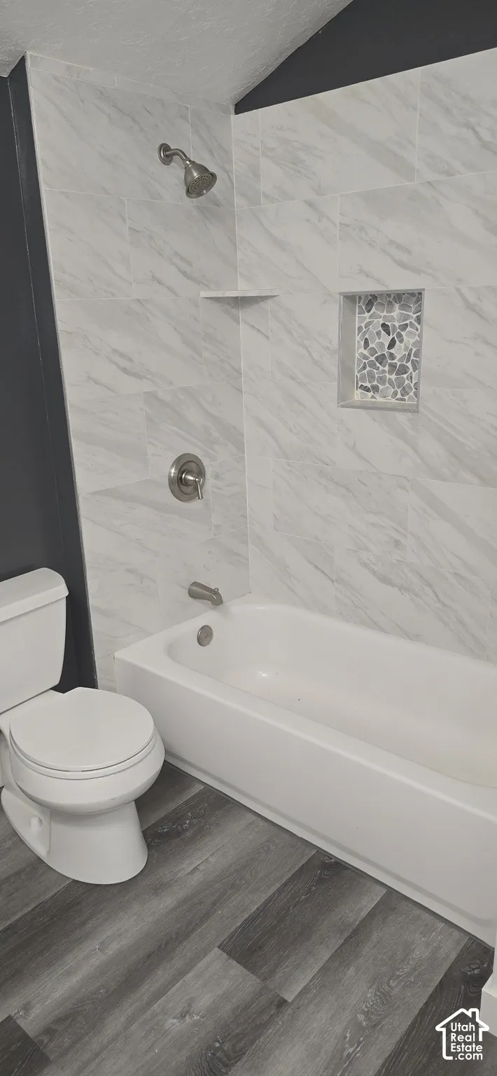 Bathroom featuring wood-finish flooring, a white porcelain tub, and a shower surround tiled in a neutral stone-patterned finish with a built-in decorative niche