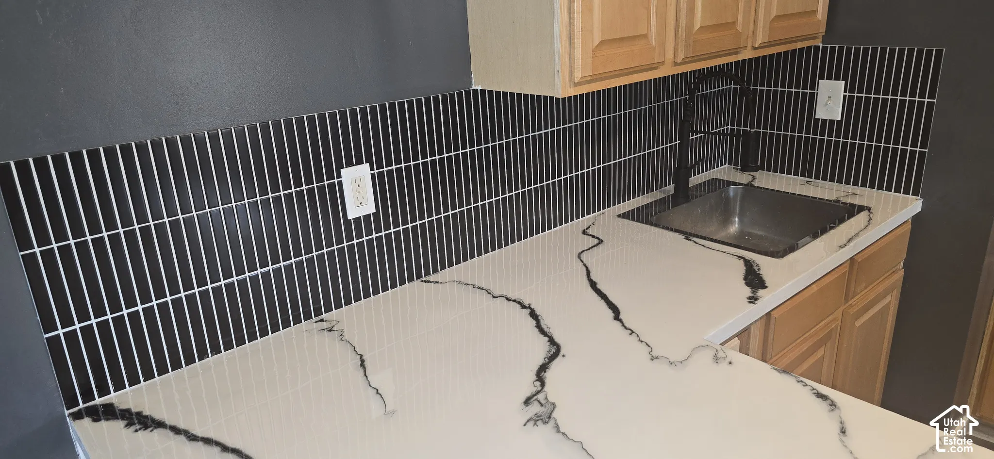Kitchen featuring white countertops with black veining, black vertical tile backsplash, stainless steel drop-in sink, matte black gooseneck faucet, and wood-finish cabinetry