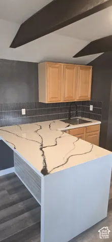 Countertop with bold veining, vertical-patterned tile backsplash, natural wood-finish cabinetry, drop-in stainless steel sink with matte black faucet, and exposed ceiling beams