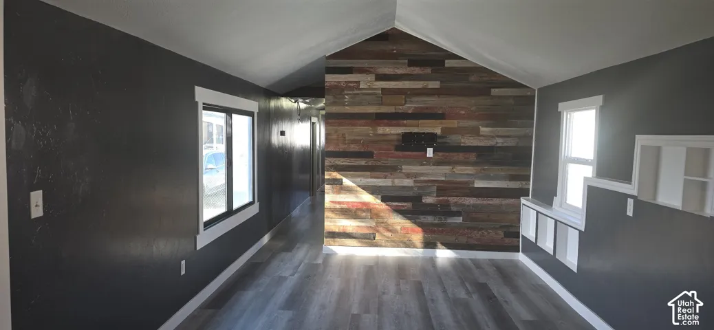 Vaulted ceiling room featuring a reclaimed wood-plank accent wall, wood-finish flooring, dark-toned interior walls, and built-in recessed shelving