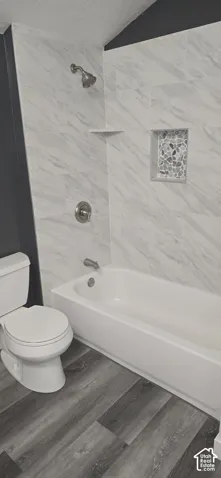 Bathroom featuring wood-finish flooring, a white porcelain tub, and a shower surround tiled in a neutral stone-patterned finish with a built-in decorative niche