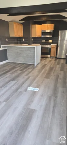 Kitchen featuring wood-finish flooring, exposed ceiling beams, stainless steel appliances, stacked stone island facade, and horizontal tile backsplash