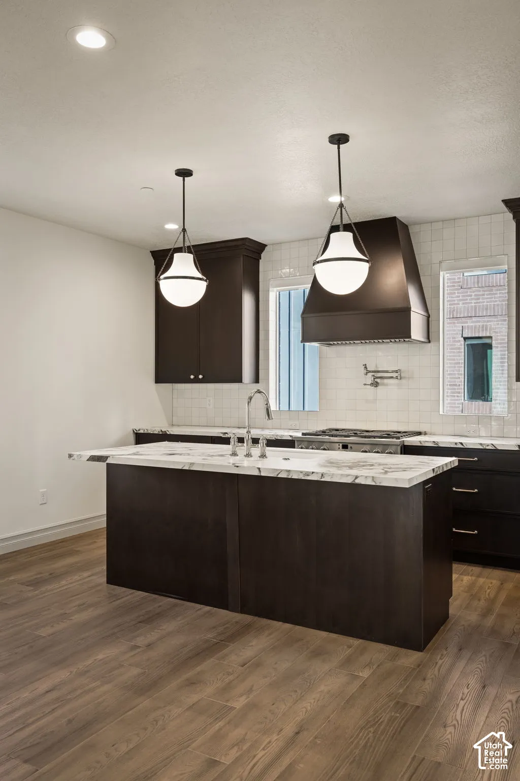Kitchen featuring dark cabinetry, a central island with stone-surface countertop, white tile backsplash, a wall-mounted pot filler, and pendant lighting
