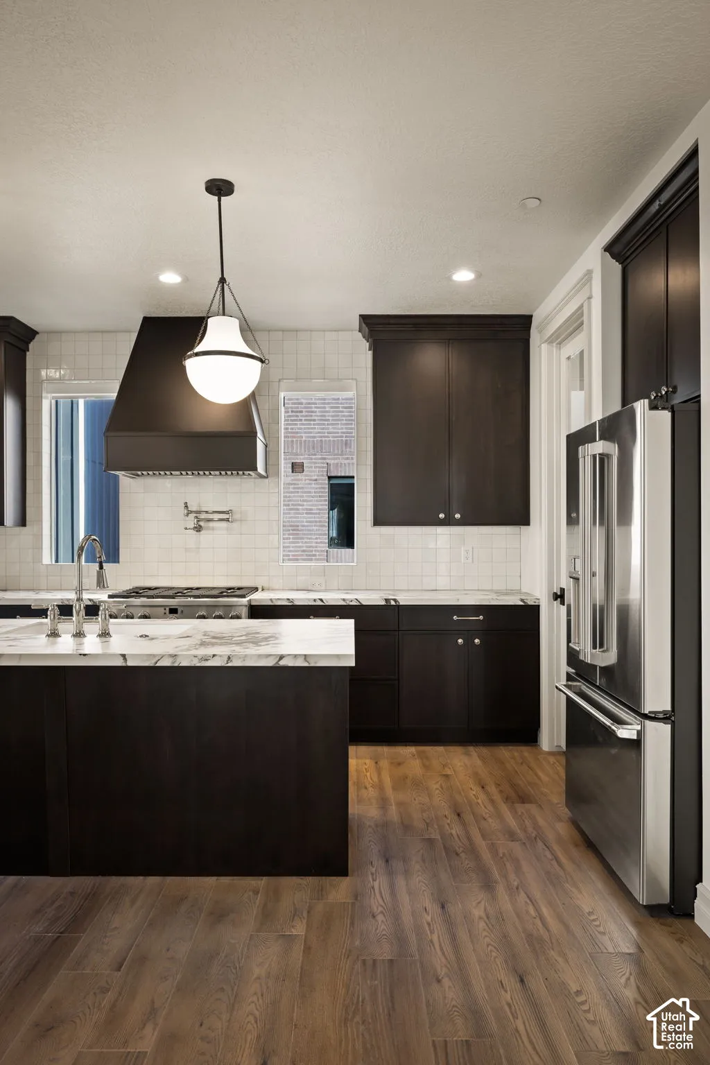 Kitchen featuring custom dark-toned cabinetry, light-toned stone countertops, a subway tile backsplash, a metal range hood, and wood-finish flooring