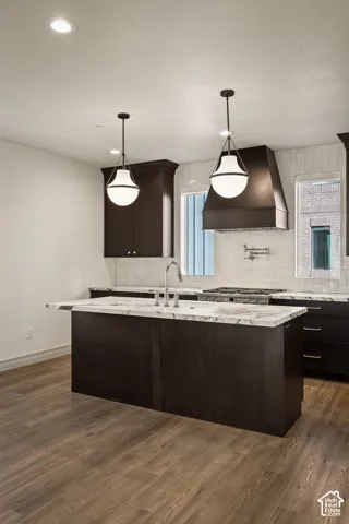 Kitchen featuring dark cabinetry, a central island with stone-surface countertop, white tile backsplash, a wall-mounted pot filler, and pendant lighting