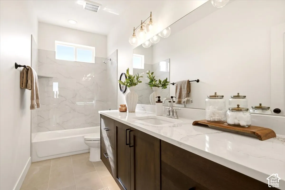 Bathroom featuring a dual-sink vanity with stone-finish countertops, dark cabinetry, full-width mirror, tile-surround shower, and neutral floor tiling
