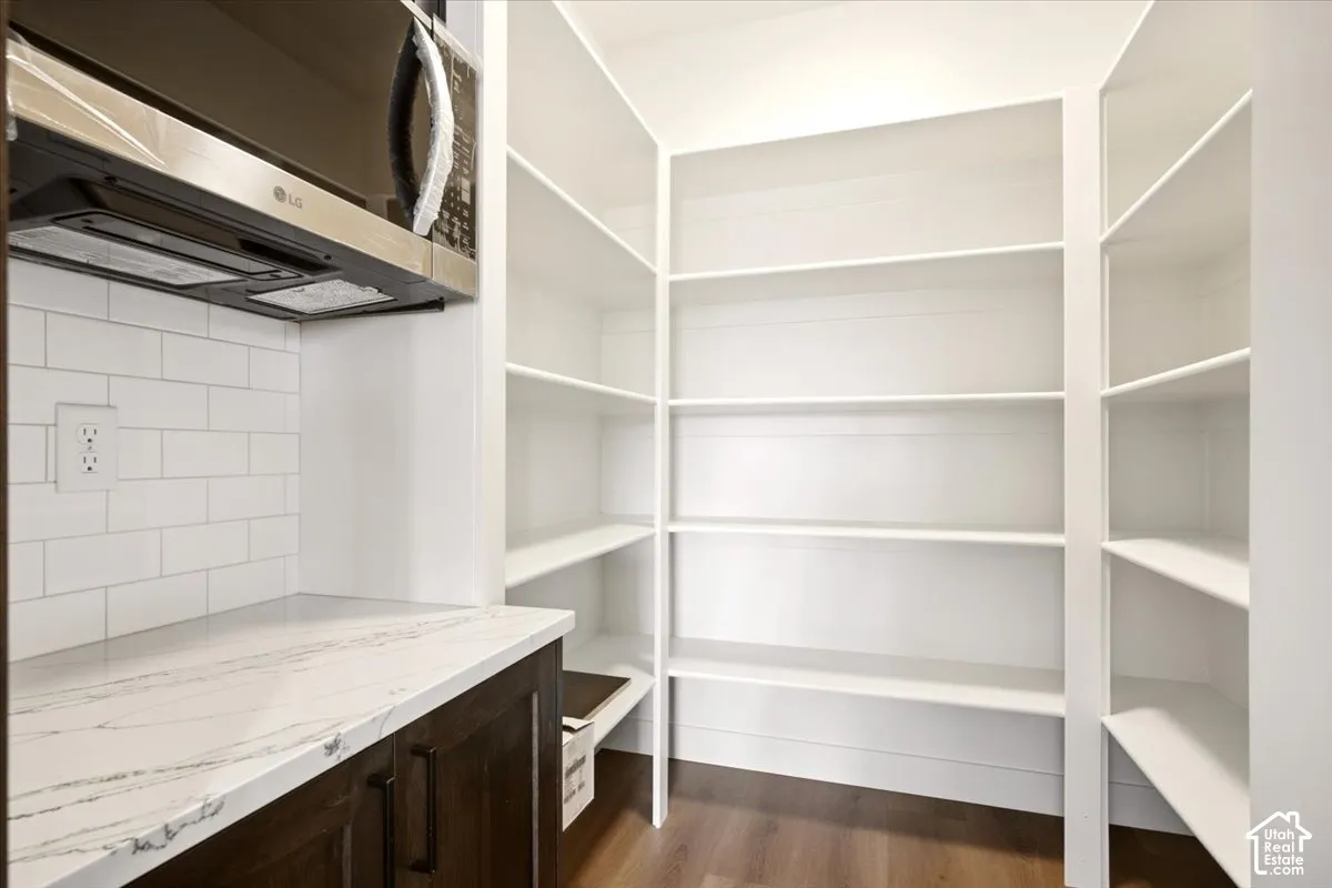 Walk-in pantry featuring white floor-to-ceiling shelving, stone-composite countertops, subway tile backsplash, dark-finish cabinetry, and wood-finish flooring