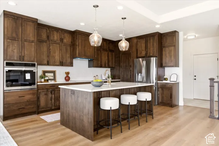 Kitchen featuring stained wood cabinetry, a central island with a solid surface countertop, stainless steel built-in appliances, a white tile backsplash, and wood-finish flooring