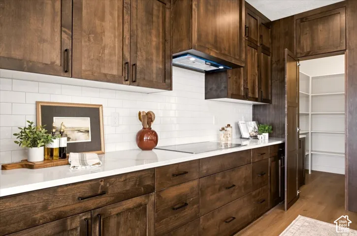 Custom dark-stained cabinetry featuring matte black hardware, white solid-surface countertops, subway tile backsplash, built-in range hood, and an attached walk-in pantry with white wire shelving