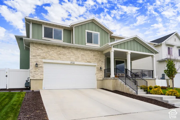 Two-story exterior featuring vertical siding, stone veneer accents, a covered front porch with columns, a concrete driveway, and a white privacy fence