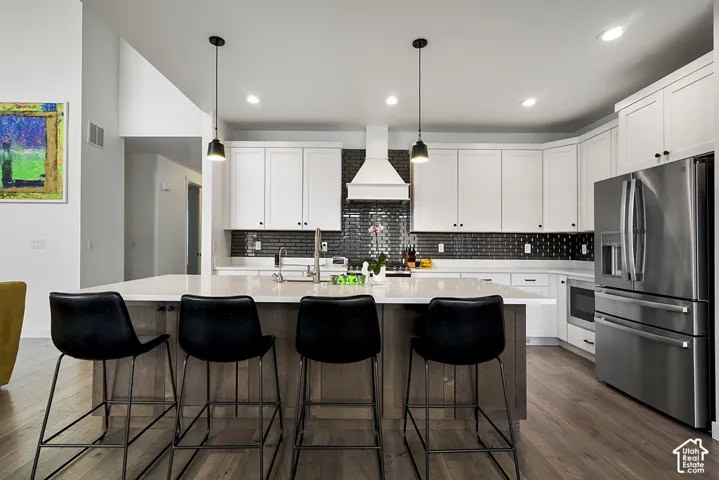 Kitchen featuring white shaker-style cabinetry, an expansive central island with stone-finish countertops, dark subway tile backsplash, stainless steel appliances, and pendant lighting