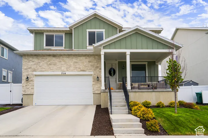 Two-story exterior featuring light-toned stone veneer, vertical board-and-batten siding, a covered front porch with a concrete stoop, and an attached two-car garage