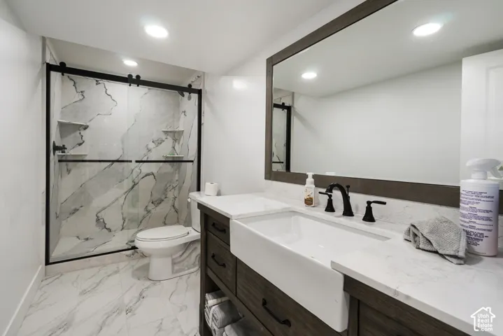 Bathroom featuring a farmhouse-style apron sink, custom wood-finish vanity with stone-look countertop, large-format stone-look tile flooring, and a walk-in shower with matte black sliding glass doors