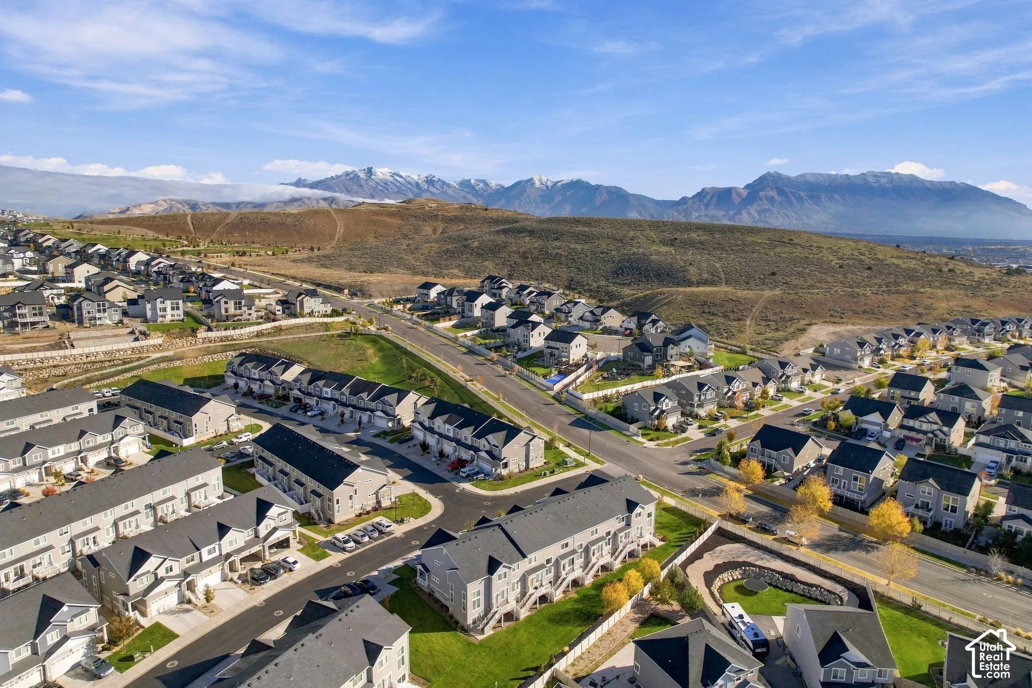 Aerial perspective of the residential community featuring multi-story dwellings, paved roadways, managed green space, and prominent mountain backdrop