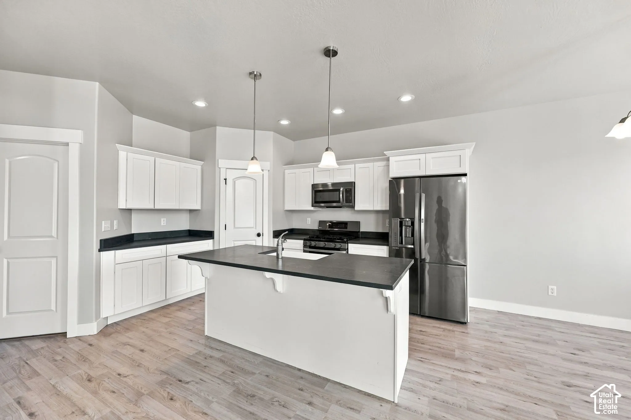 Kitchen featuring a central island with a sink, white cabinetry, dark countertops, wood-finish flooring, and stainless steel appliances