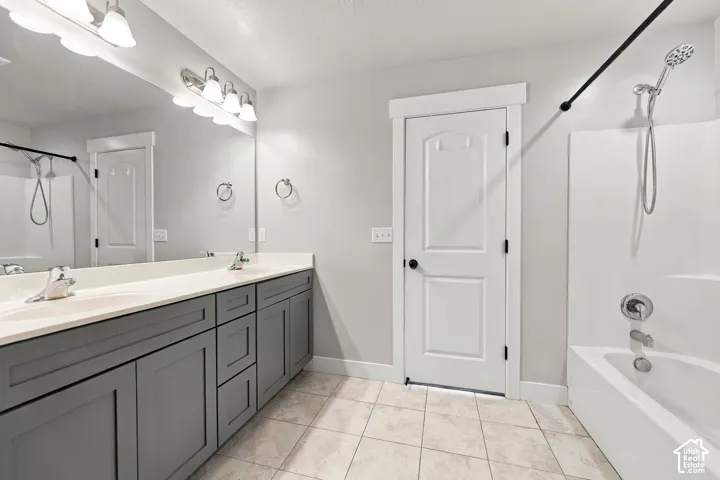 Bathroom featuring a dual-sink vanity with gray cabinetry, solid-surface countertop, full-width vanity mirror, tile flooring, and a tub-shower combination with a handheld showerhead