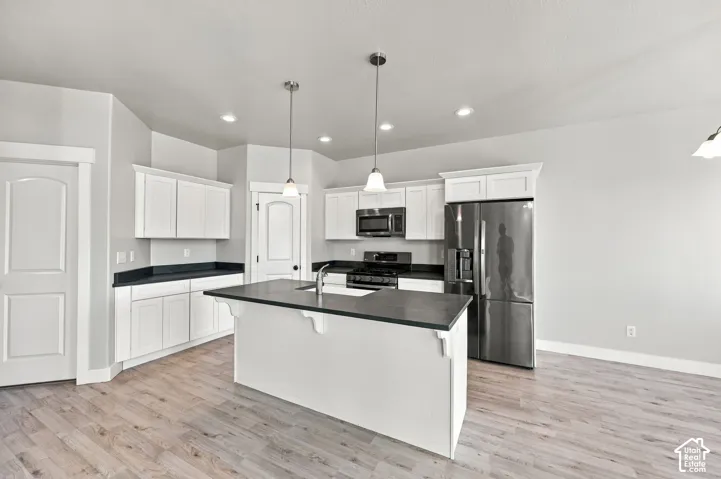 Kitchen featuring a central island with a sink, white cabinetry, dark countertops, wood-finish flooring, and stainless steel appliances