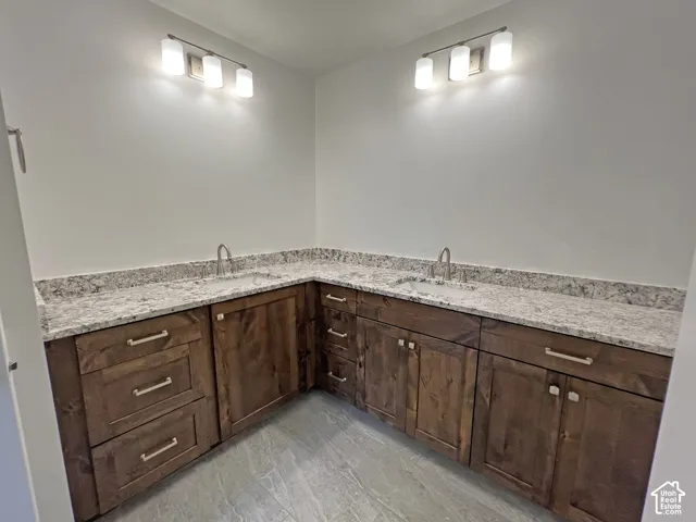 Double vanity featuring stained wood cabinetry, stone-patterned countertops with integrated sinks, dual chrome faucets, and two modern three-light fixtures