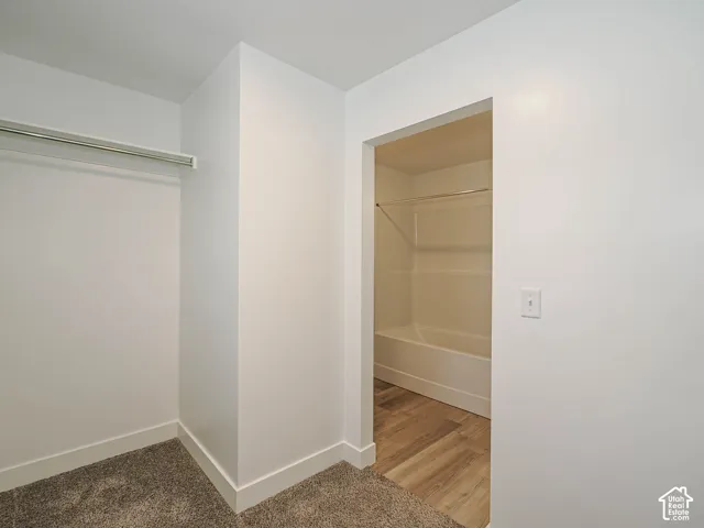 Walk-in storage space with metal hanging rod and neutral carpeting, featuring an interior bathroom with wood-finish flooring and a white tub-shower surround