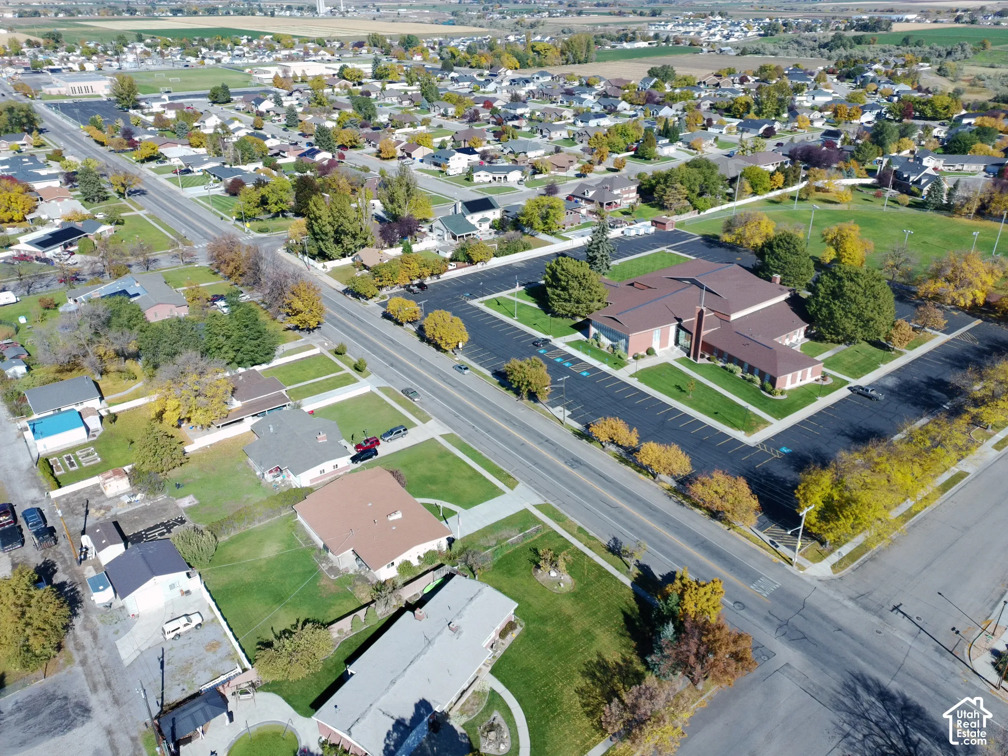 Aerial perspective of a suburban neighborhood featuring paved street infrastructure, residential lots with lawn landscaping, mature trees, and an institutional property with a large parking area