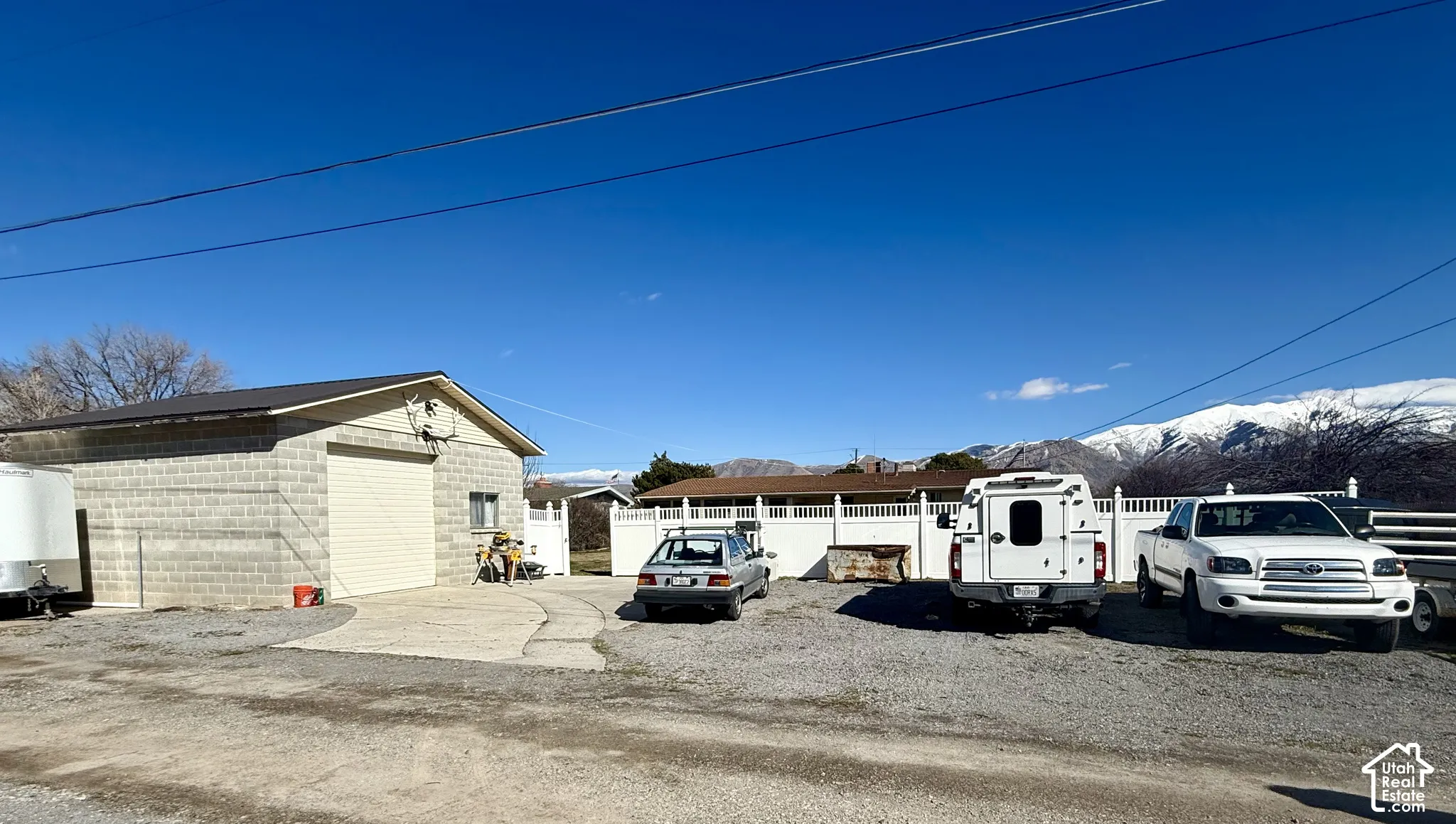 Detached concrete masonry structure featuring a roll-up bay door, concrete apron, and white vinyl perimeter fencing, set against a mountain backdrop