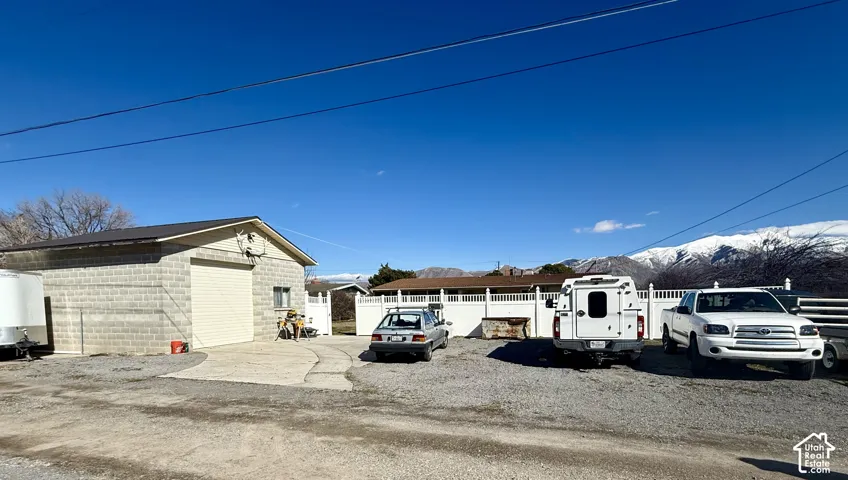 Detached concrete masonry structure featuring a roll-up bay door, concrete apron, and white vinyl perimeter fencing, set against a mountain backdrop