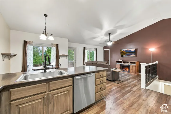 Open-concept kitchen and living area featuring vaulted ceilings, wood-finish flooring, stainless steel dishwasher, undermount kitchen sink with high-arc faucet, and a built-in media console with electric fireplace
