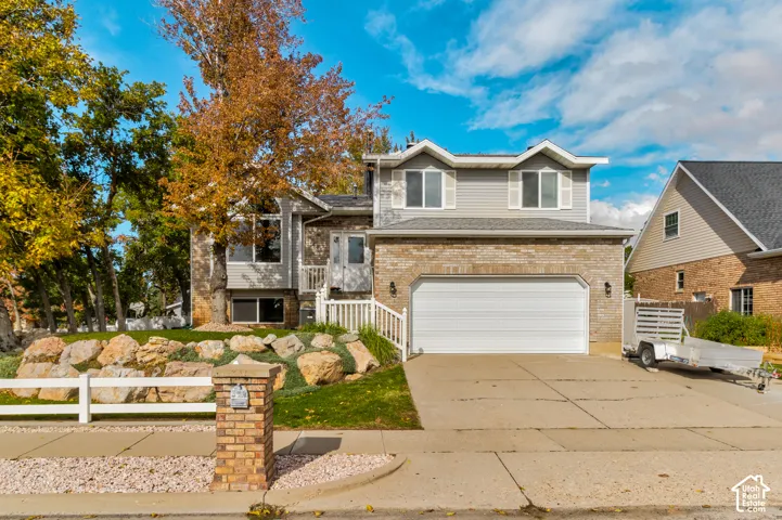 Split-level exterior featuring light brick and horizontal siding, an attached two-car garage, tiered stone landscaping, and a white picket fence