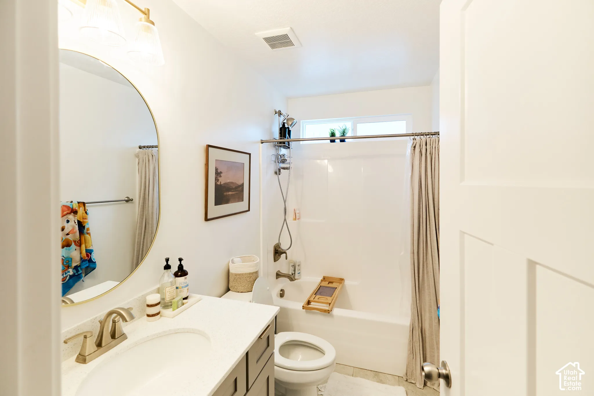 Bathroom featuring a vanity with a white countertop and oval undermount sink, brass-toned faucet, circular mirror with a metallic frame, and a tub-shower combination with a transom window
