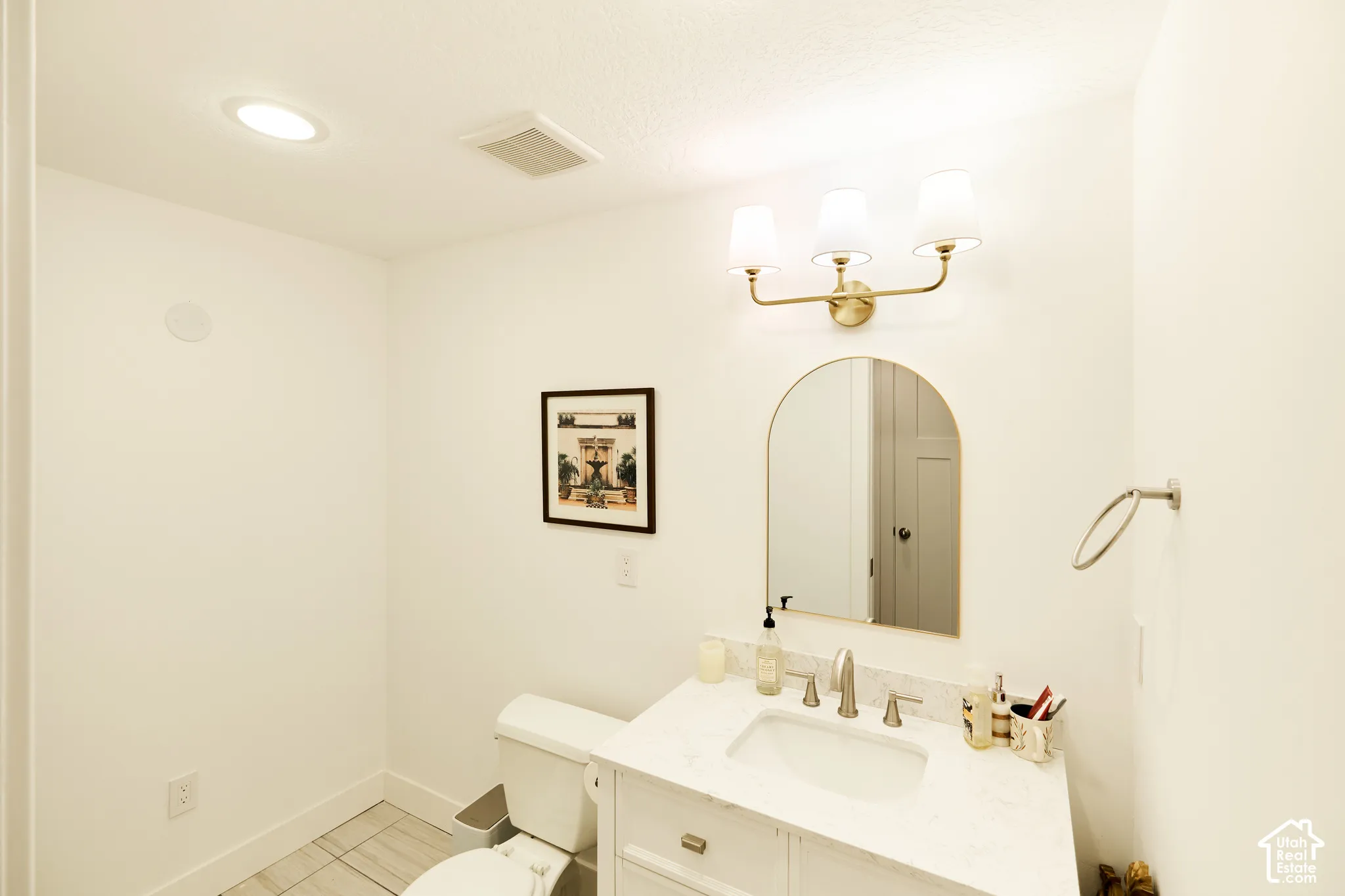 Bathroom featuring a vanity with a stone-finish countertop, an arched-top mirror, brass-finish three-light fixture, and ceramic tile flooring
