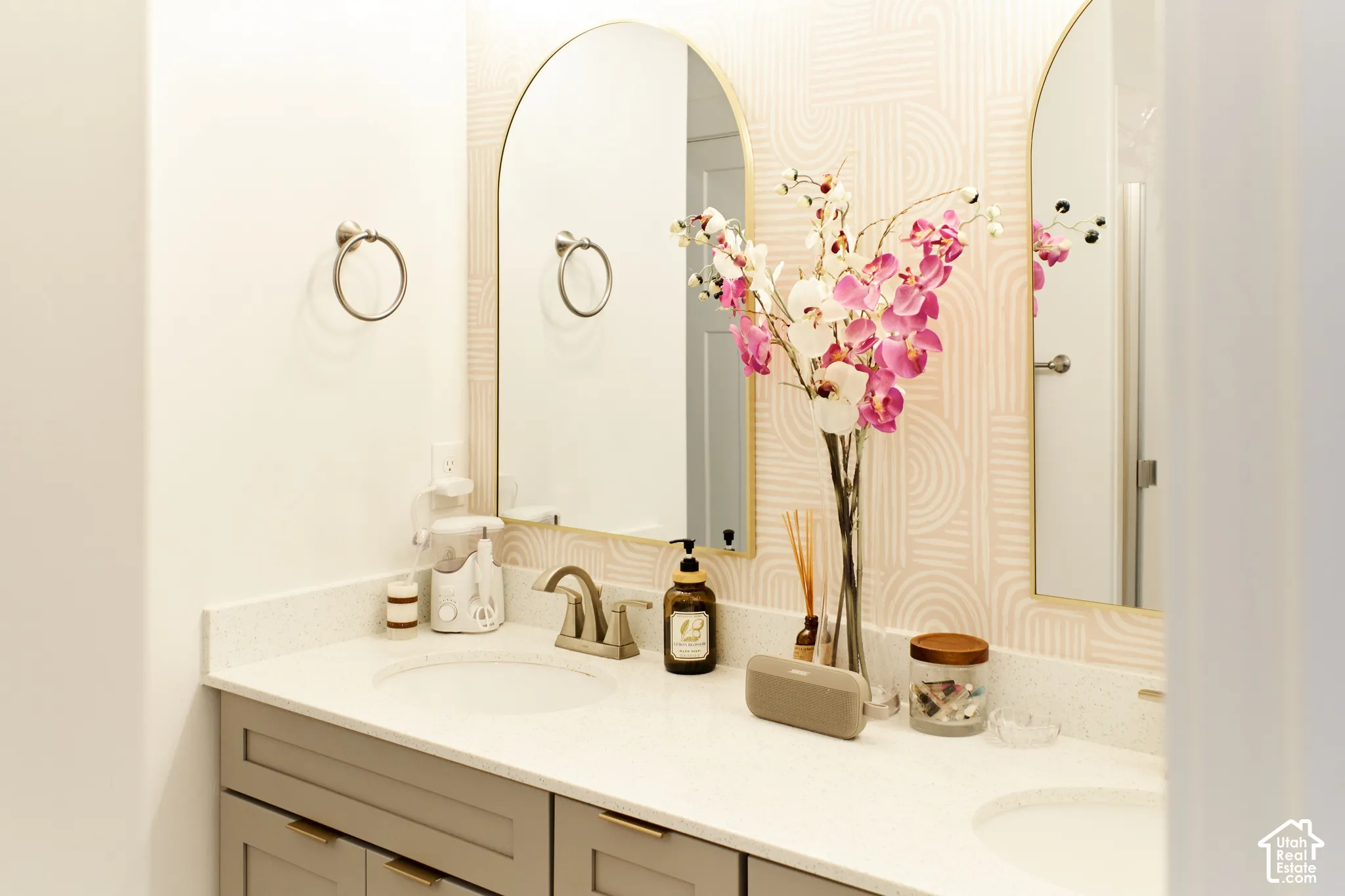 Dual-sink vanity with a solid-surface countertop, arched gold-rimmed mirrors, patterned wallpaper, and gray shaker-style cabinetry with metallic hardware