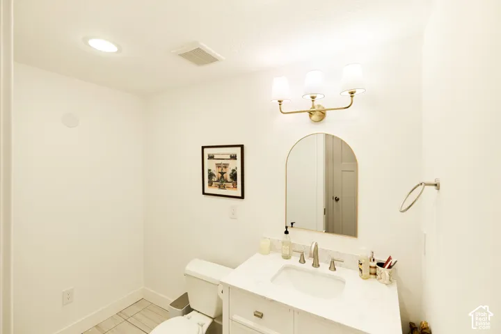 Bathroom featuring a vanity with a stone-finish countertop, an arched-top mirror, brass-finish three-light fixture, and ceramic tile flooring