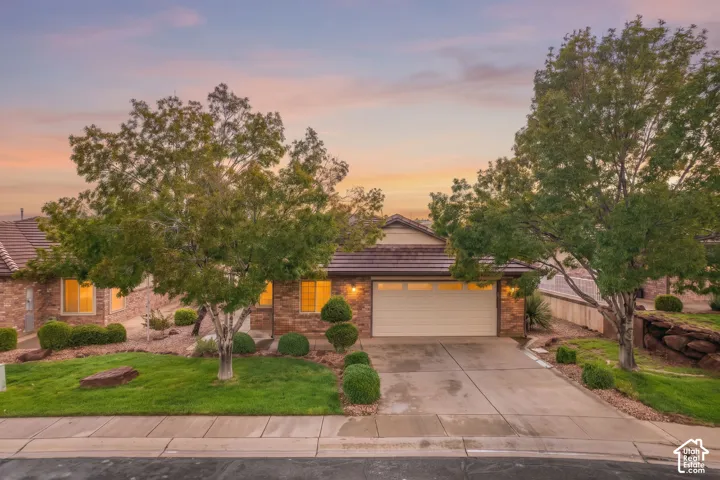 Single-story exterior featuring masonry stone veneer, a tiled roof, concrete driveway, and professional landscaping with mature trees and shaped shrubs