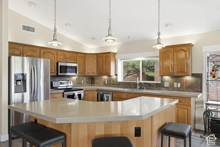 Kitchen featuring wood cabinetry, solid-surface countertops, stone tile backsplash, stainless steel appliances, and three metal pendant lights
