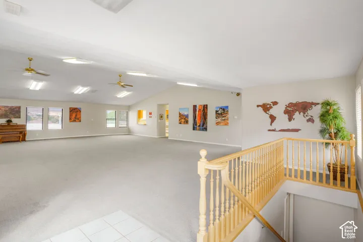Expansive open-concept room featuring vaulted ceilings, neutral wall-to-wall carpeting, multiple ceiling fans, and wood-railed staircase balusters