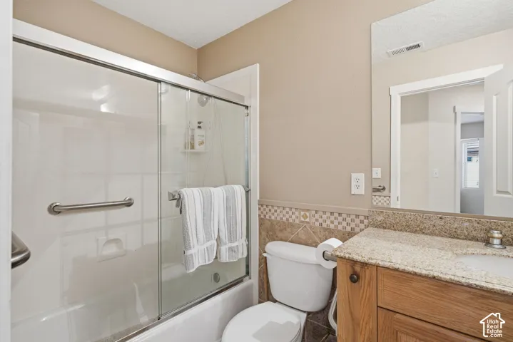 Bathroom featuring a tub-shower combination with glass sliding doors and a metal grab bar, vanity with stone-finish countertop, decorative tile backsplash, and wall-mounted mirror